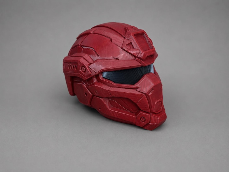 May include: A deep red tactical helmet featuring a segmented, multi-piece construction with a dark, recessed visor and a prominent rectangular mount on the forehead. The surface displays a textured finish with visible panel lines and geometric details.