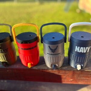 May include: Four insulated water coolers with handles, each featuring a different military branch: Army, Marines, Air Force, and Navy. The coolers are in olive green, red, gray, and navy blue, with matching handles and text. Each has a spigot for dispensing liquids.