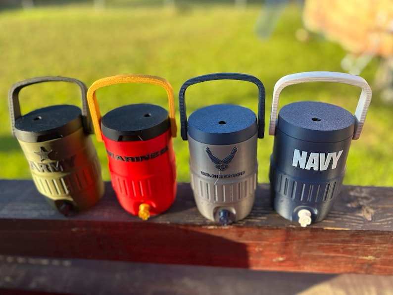 May include: Four insulated water coolers with handles, each featuring a different military branch: Army, Marines, Air Force, and Navy. The coolers are in olive green, red, gray, and navy blue, with matching handles and text. Each has a spigot for dispensing liquids.