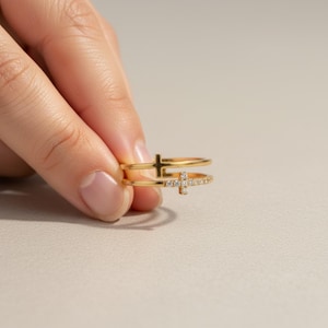 May include: A gold double-band ring featuring two distinct cross motifs. The top band displays a plain polished gold cross, while the bottom band features a cross encrusted with small, clear, sparkling stones along its surface.