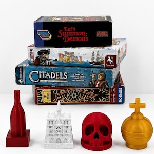 May include: A stack of four board games titled Let's Summon Demons, Port Royal, Citadels, and The Crew, accompanied by four 3D-printed game tokens: a red bottle, a white cathedral, a red skull, and a gold orb with a cross on top.