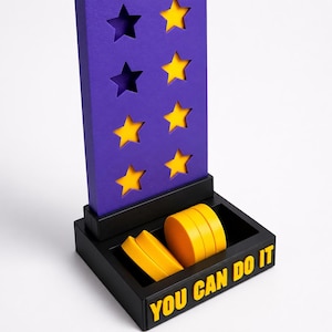 May include: A purple vertical reward board with eight star-shaped cutouts, some filled with yellow inserts. The board sits in a black base containing several yellow circular tokens. The front of the base features the yellow text YOU CAN DO IT.