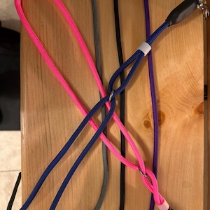 May include: A collection of dog leashes in various colors, including pink, blue, gray, black, and purple. Each leash is made of a woven rope material and features a metal clasp for attachment. The leashes are arranged on a wooden surface.