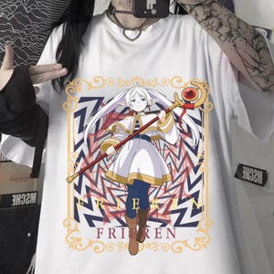 Pode incluir: Camiseta branca com um gr&aacute;fico de anime colorido de um personagem segurando uma bengala. O gr&aacute;fico inclui o texto "Frieren" em uma fonte estilizada, em um fundo geom&eacute;trico. O personagem &eacute; retratado com um vestido branco e botas marrons.