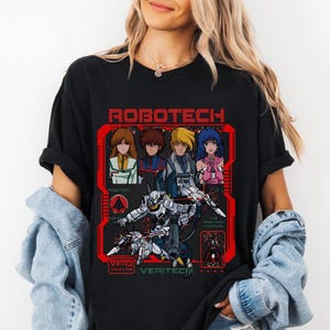May include: Black t-shirt featuring a red graphic with the word "ROBOTECH" at the top. The graphic includes anime-style characters and a robot. The text "VF-1J VALKYRIE VERITECH" is also present. The shirt is worn by a person.