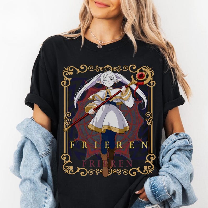 Pode incluir: Camiseta preta com um gr&aacute;fico detalhado de um personagem de anime segurando uma bengala. O personagem est&aacute; vestido com roupas brancas e azuis, emoldurado em uma moldura decorativa com a palavra "FRIEREN" em letras douradas. Tema de fantasia.