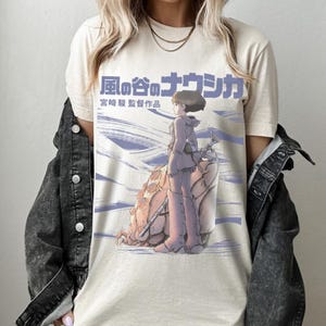 Peut inclure: T-shirt cr&egrave;me avec une illustration d'anime japonais. Le motif comprend un personnage en tenue violette, debout &agrave; c&ocirc;t&eacute; d'une grande cr&eacute;ature, avec du texte japonais au-dessus. Le t-shirt a un col rond classique et des manches courtes.
