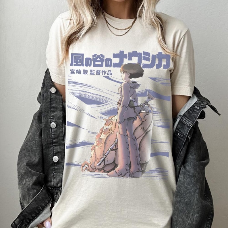Peut inclure: T-shirt cr&egrave;me avec une illustration d'anime japonais. Le motif comprend un personnage en tenue violette, debout &agrave; c&ocirc;t&eacute; d'une grande cr&eacute;ature, avec du texte japonais au-dessus. Le t-shirt a un col rond classique et des manches courtes.