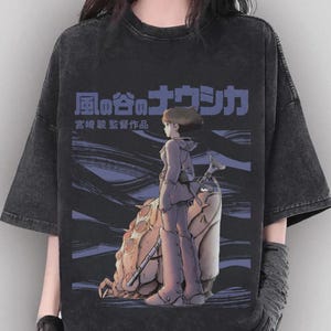 Peut inclure: T-shirt gris fonc&eacute; avec un graphisme du film d'animation "Nausica&auml; de la Vall&eacute;e du Vent". Le motif comprend du texte japonais et une illustration d'un personnage en tenue violette. Le t-shirt a un aspect vieilli.