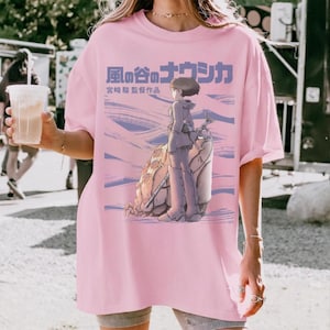 Peut inclure: T-shirt rose clair avec un motif d'anime japonais. Le motif comprend du texte japonais et une illustration d'un personnage avec une cr&eacute;ature et un paysage dans des tons violets et bleus.