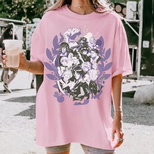 Pu&ograve; includere: T-shirt rosa chiaro con un disegno di personaggi anime in un cerchio. La grafica utilizza tonalit&agrave; di nero, bianco e viola, incorniciata da foglie. La maglietta ha maniche corte e una vestibilit&agrave; ampia.