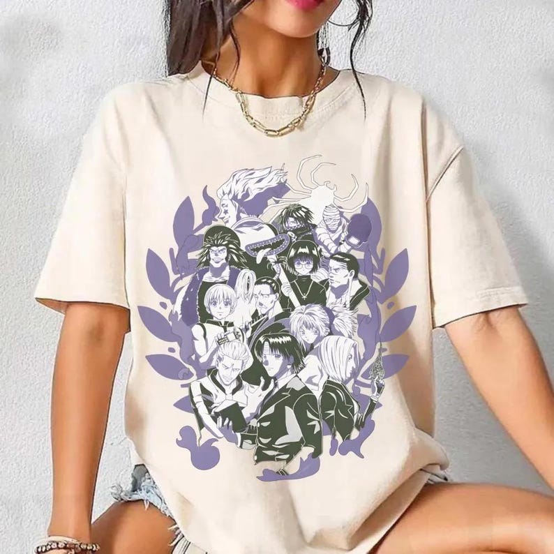 Pu&ograve; includere: T-shirt beige con una grafica di personaggi anime in bianco, nero e grigio, incorniciata da un design a foglie viola. L'immagine &egrave; stampata sulla parte anteriore della maglietta.