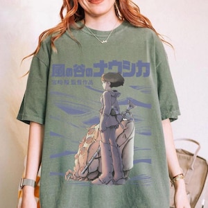 Peut inclure: T-shirt vert avec un graphisme d'anime japonais. Le motif comprend un personnage avec une arme, une grande cr&eacute;ature ressemblant &agrave; un insecte et du texte japonais. Le t-shirt a un aspect vintage et d&eacute;lav&eacute;.