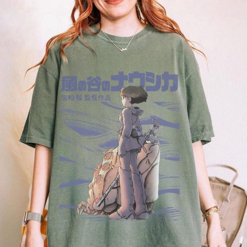 Peut inclure: T-shirt vert avec un graphisme d'anime japonais. Le motif comprend un personnage avec une arme, une grande cr&eacute;ature ressemblant &agrave; un insecte et du texte japonais. Le t-shirt a un aspect vintage et d&eacute;lav&eacute;.
