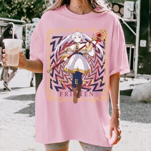 Pode incluir: T-shirt rosa claro com um gr&aacute;fico de um personagem de anime segurando uma bengala. O personagem est&aacute; vestido com uma roupa branca e dourada e tem cabelo comprido e branco. A palavra "FRIEREN" est&aacute; impressa abaixo do personagem na camiseta.