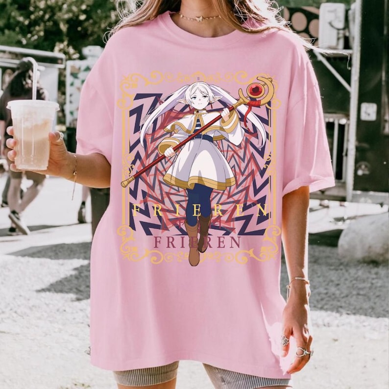 Pode incluir: T-shirt rosa claro com um gr&aacute;fico de um personagem de anime segurando uma bengala. O personagem est&aacute; vestido com uma roupa branca e dourada e tem cabelo comprido e branco. A palavra "FRIEREN" est&aacute; impressa abaixo do personagem na camiseta.