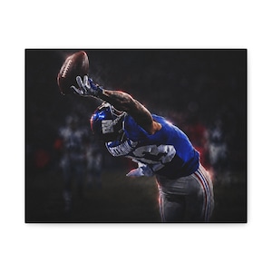 May include: A football player in a blue jersey with the number 13 and white trousers reaching upwards with one hand to catch a brown football. The player wears a blue helmet and white gloves, captured in a dynamic motion against a dark background.