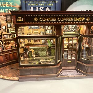 May include: A miniature Corner Coffee Shop model with detailed interior and exterior features. The model includes a bookshelf, tables, chairs, and decorative elements. The shop's facade features the text "CORNER COFFEE SHOP" in gold lettering.