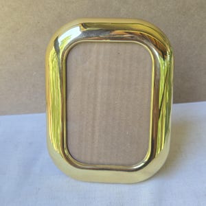 Op de afbeelding: Een verticale, rechthoekige fotolijst met afgeronde hoeken en een gepolijste, reflecterende goudkleurige metalen afwerking. De lijst heeft een brede, gladde rand rondom een centrale opening die een effen, lichtbruin kartonnen inzetstuk bevat.