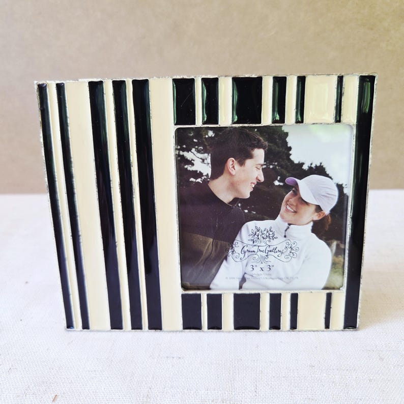 May include: A decorative photo frame with a black and cream striped pattern. The frame holds a 3" x 3" photo of a couple. The frame is made of metal and has a glossy finish. The frame is a decorative item.