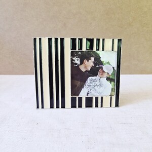 May include: A black and cream striped photo frame with a 3x3 inch photo. The frame has a rectangular shape with vertical stripes. The photo shows two people smiling at each other. The frame is on a white surface.