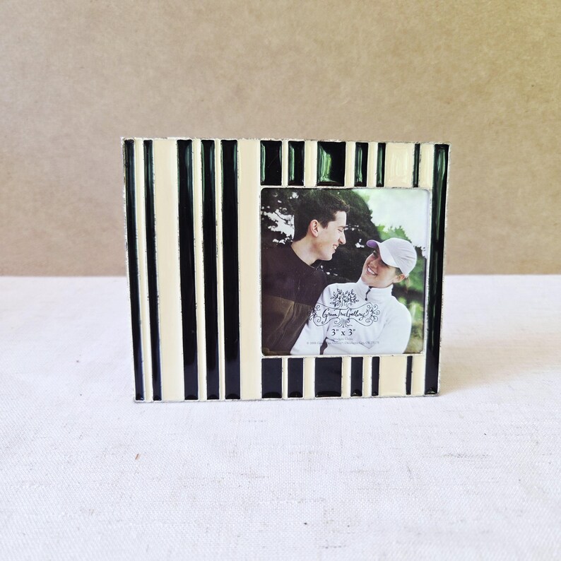 May include: A black and cream striped photo frame with a 3x3 inch photo. The frame has a rectangular shape with vertical stripes. The photo shows two people smiling at each other. The frame is on a white surface.