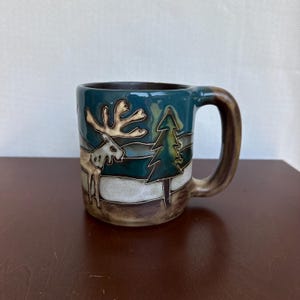 Può includere: Una tazza in ceramica con smalto color verde acqua e marrone, caratterizzata da un design in rilievo raffigurante un alce accanto a un pino. La tazza presenta un manico robusto e una finitura liscia e lucida con un paesaggio innevato.
