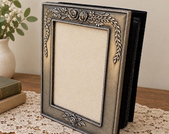 May include: A rectangular photo frame with an antique bronze finish, featuring embossed rose and vine motifs along the top and bottom edges. The frame has a rope-textured inner border and a plain beige insert, standing upright on a flat surface.