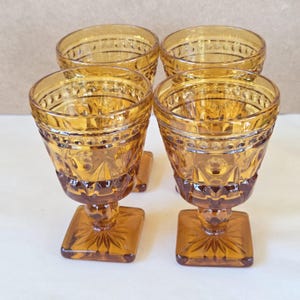 May include: Four amber-colored vintage goblets. Each goblet features a square base with a starburst design, a short stem, and a patterned bowl. The rims have a dotted design. The glasses are made of glass.