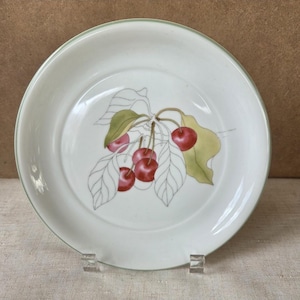 May include: A white ceramic plate with a green rim and a cherry design. The design features red cherries and green leaves with a light brown stem. The plate is round and has a slightly raised edge.