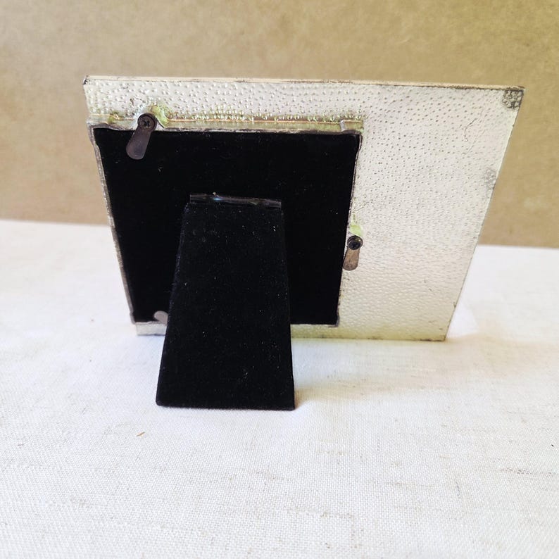 May include: A small, rectangular picture frame with a textured, off-white finish. The back of the frame is black, with a black velvet easel stand. The frame is angled, showing the back and side.