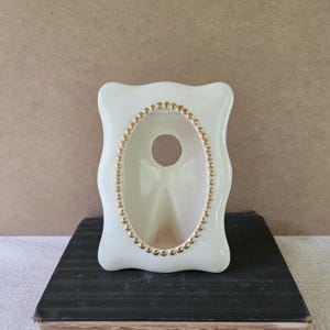 Puede incluir: Un calentador de aceite de cerámica blanca brillante con un marco rectangular de bordes ondulados y una abertura ovalada central. El óvalo está bordeado por una fila decorativa de pequeñas cuentas doradas en relieve. Incluye un recorte circular.
