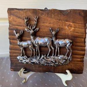 Peut inclure: Une plaque murale rectangulaire composée d'une planche en bois brun foncé à bord naturel, ornée d'un relief en métal argenté représentant un cerf debout entre deux biches plus petites. Les figures métalliques sont texturées de taches sombres.