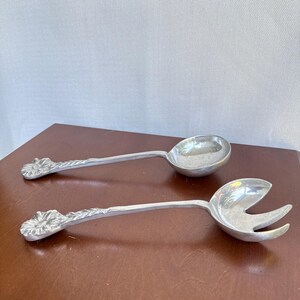 Può includere: Una coppia di posate da insalata in metallo color argento con finitura lucida. Ogni manico è decorato con un motivo floreale scolpito all'estremità. Un utensile è un cucchiaio rotondo pieno, l'altro è un cucchiaio abbinato a due rebbi.