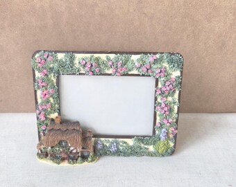 May include: A rectangular picture frame with a cottage scene. The frame is decorated with pink roses and green foliage. The cottage has a thatched roof and is surrounded by greenery. The frame is made of a cream-colored material.