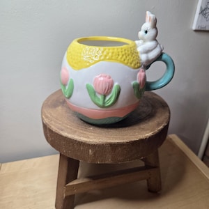 May include: A ceramic Easter-themed mug with a white, yellow, and pink egg design, featuring a bunny handle and tulip accents. The mug is placed on a small, round wooden stool, showcasing a rustic aesthetic. The mug's handle is a light blue color.
