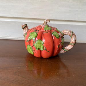 Puede incluir: Una tetera de cerámica con forma de calabaza de color naranja brillante y acabado esmaltado. La tetera presenta hojas y enredaderas verdes en relieve sobre el cuerpo, con un asa y un pico diseñados para parecer ramas de árbol marrones.