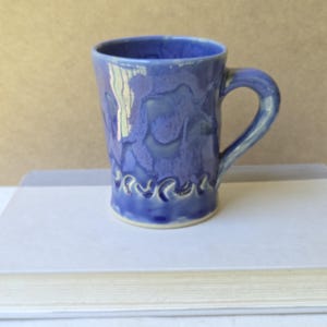 Puede incluir: Una taza de cerámica con un esmalte brillante en varios tonos de azul. La superficie muestra un patrón moteado similar a la acuarela con una banda texturizada en forma de ondas de color azul oscuro que rodea la parte inferior del recipiente.