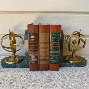 May include: A set of three Reader's Digest Condensed Books positioned between two brass armillary sphere bookends on a dark green marble base. The books feature brown and red spines with gold lettering, displaying titles such as The White Dawn and Airport.