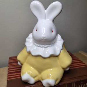 May include: A ceramic bunny figurine with a white head and ears, red eyes, and a white ruffled collar. The bunny wears a yellow outfit. The figurine is sitting on a stack of books. This decorative item is suitable for Easter.