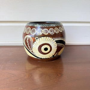 Könnte beinhalten: Ein bauchiger Keramiktopf mit dunkelbrauner Grundfarbe und handgemalten cremefarbenen sowie schwarzen Verzierungen. Das Design zeigt ein markantes zentrales Blumenmotiv, geschwungene Spiralen am oberen Rand und feine lineare Details.