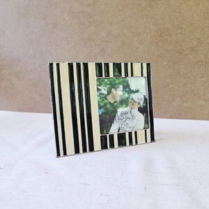 May include: A square photo frame with a black, cream, and green striped design. The frame holds a photograph of two people. The photo frame is 3x3 inches.