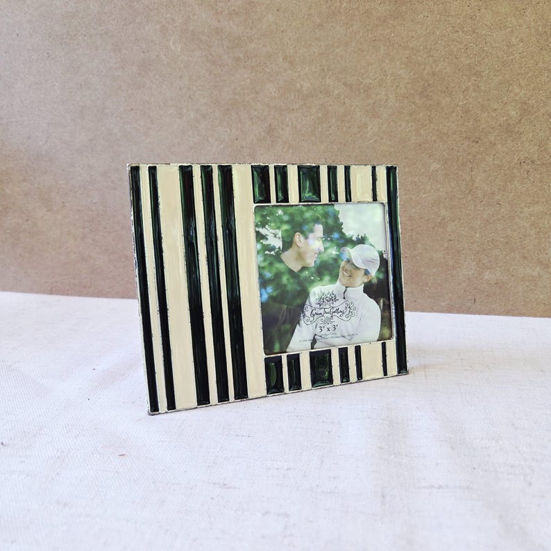 May include: A square photo frame with a black, cream, and green striped design. The frame holds a photograph of two people. The photo frame is 3x3 inches.