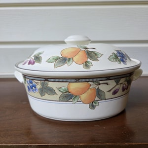 May include: A round white ceramic casserole dish with a matching lid, featuring a decorative band of painted yellow pears, green leaves, and clusters of blue and purple grapes. The dish has two small side handles and a central knob on the lid.