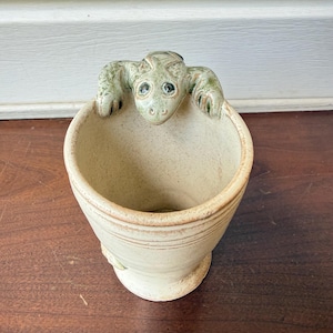 May include: A beige ceramic mug featuring a sculpted green frog figure perched on the rim. The mug has a speckled texture, a flared base, and horizontal grooved lines around the exterior. The frog has dark eyes and textured skin details.