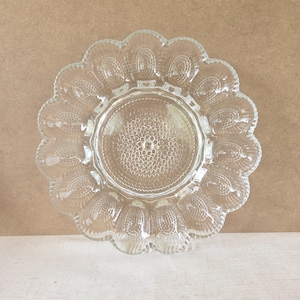 Könnte beinhalten: Klarer Glas-Servierteller mit gewelltem Rand. Die Mitte des Tellers hat ein strukturiertes, kreisförmiges Muster, und der äußere Rand weist ein sich wiederholendes Design auf. Der Teller ist für Eier konzipiert.