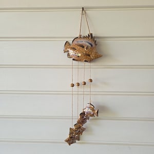 May include: A hanging wall decoration featuring a large ceramic fish at the top, connected by four thin jute strings to a smaller fish at the bottom. The fish are glazed in shades of brown and tan with a speckled finish. Four small wooden beads are strung along the cords.