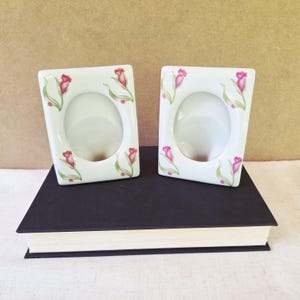 May include: Two white ceramic picture frames with oval openings, decorated with pink and red tulip designs and green leaves. The frames are rectangular with rounded corners and sit on a dark book.