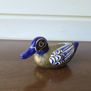 May include: A duck figurine featuring a metallic brass-toned body with a textured scale pattern. The head is painted in a deep glossy blue with white accents around the eye, and the wings display a detailed blue and white feather pattern.