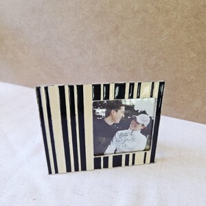 May include: A black and cream striped photo frame with a photograph of two people. The frame has a rectangular shape with a slightly angled design. The photograph is in black and white.